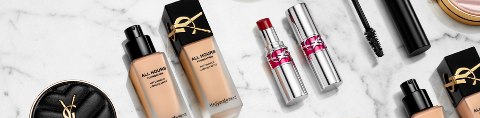 Make-up-Produkte von Yves Saint Laurent auf einer weißen Marmoroberfläche, darunter zwei Flaschen flüssige Foundation, zwei silberne Lippenstifte, eine Mascara und ein schwarzes Puderdöschen mit goldenem YSL-Logo.