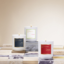 Drei Duftkerzen von Maison Francis Kurkdjian Paris stehen brennend auf einer gemusterten Fläche aus Marmor- und Steinplatten. Die weißen Kerzengläser tragen Etiketten in Dunkelblau, Neongrün und Rot mit goldener bzw. weißer Schrift.