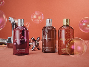 Drei Duschgelflaschen von Molton Brown in verschiedenen Farben – Pink, Braun und Bernstein – stehen nebeneinander auf einer orangeroten Fläche. Um sie herum schweben transparente Seifenblasen. Im Hintergrund befinden sich metallische Wasserhähne und Armaturen, die an ein Spa oder Badezimmer erinnern. Die Szene wirkt verspielt, luxuriös und pflegend.