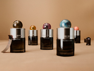 Fünf stilvolle Parfumflakons von Molton Brown mit glänzenden silbernen Verschlüssen und kugelförmigen, bunt gemusterten Kappen stehen auf einem beigen Untergrund. Die farbigen Kugeln in Grün, Orange, Violett, Blau und Rosa verleihen jedem Flakon eine individuelle Note. Im Vordergrund liegt ein Filmstreifen, im Hintergrund ein kleiner Projektor. Die Szene wirkt kreativ, edel und modern.