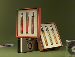 Zwei geöffnete Geschenkboxen mit je vier schmalen Parfum-Miniaturen von Molton Brown stehen vor olivgrünem Hintergrund. Eine Box ist auf einer silbernen Kompaktkamera platziert, die andere liegt leicht angewinkelt daneben. Im Vordergrund sind eine Audiokassette und eine transparente Hülle zu sehen. Die Szene wirkt nostalgisch und hochwertig.