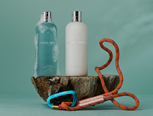 Zwei Haarpflegeprodukte von Molton Brown – eine hellblaue Shampoo-Flasche mit Schaum und eine weiße Conditioner-Flasche – stehen auf einem Stück Baumrinde vor mintgrünem Hintergrund. Davor liegt ein orangefarbenes Kletterseil mit blauem Karabiner. Die Szene vermittelt Frische, Pflege und Abenteuerlust.