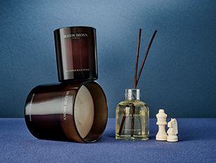 Zwei dunkelbraune Duftkerzen von Molton Brown, eine stehend, die andere liegend, zusammen mit einem Raumduft-Diffuser mit Holzstäbchen. Die Szene ist auf einer blauen Oberfläche mit blauem Hintergrund arrangiert. Rechts stehen drei weiße Schachfiguren – König, Dame und Springer – als dekoratives Element. Das Bild vermittelt Eleganz und Gemütlichkeit.